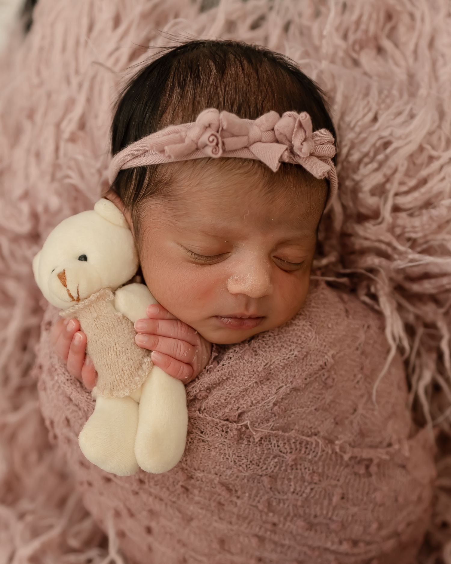 Sleeping newborn wrapped in pink fabric holding small plush bunny toy with floral headband.