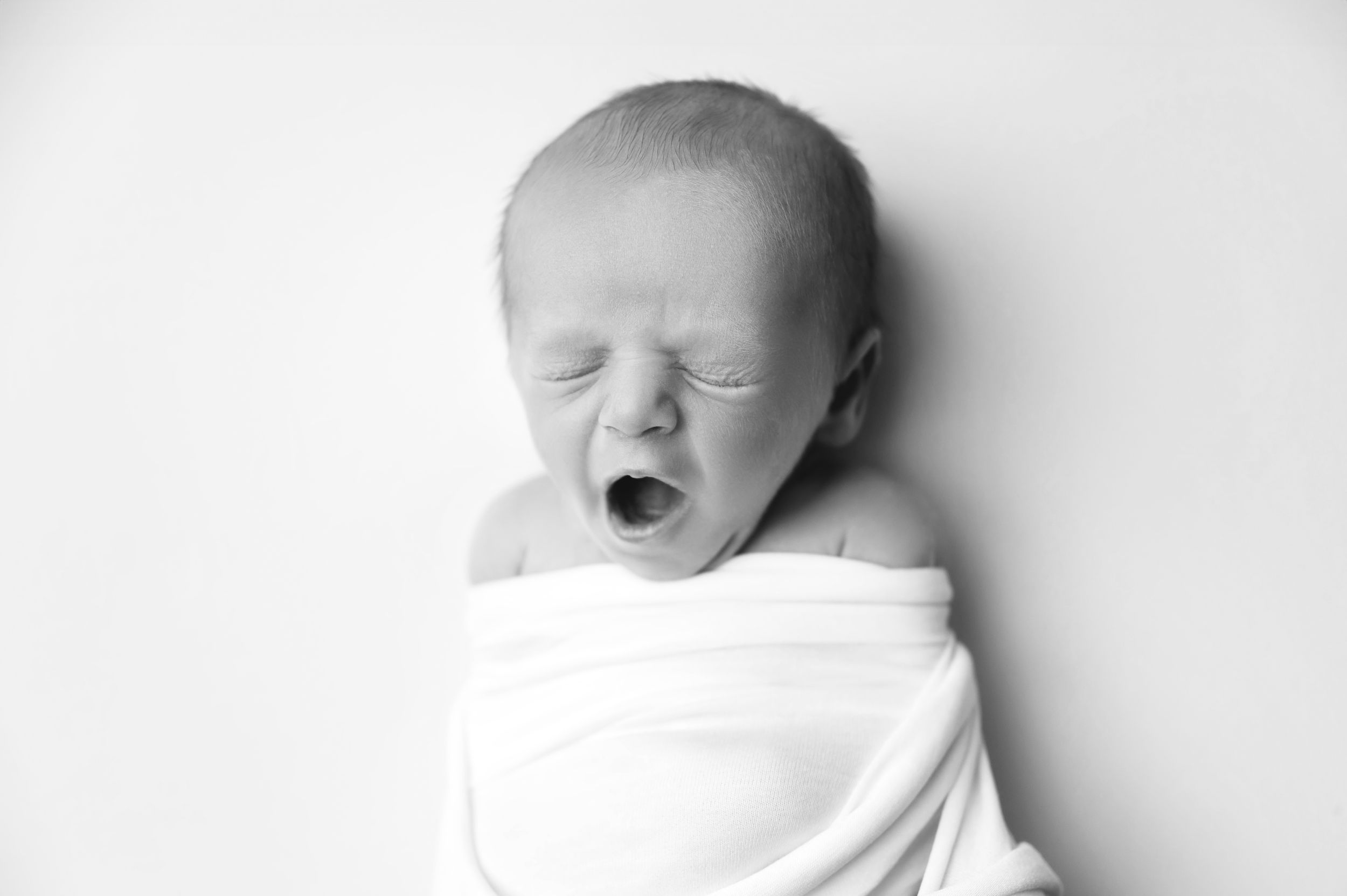 A black and white photo of a yawning newborn baby wrapped in a white blanket against a light background. Fresno Newborn Photographer