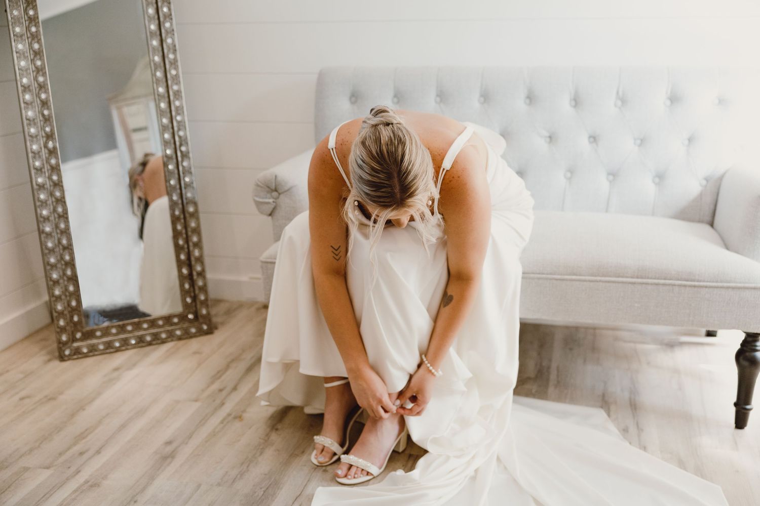 A bride in a white dress fastens shoes while sitting on an elegant tufted sofa next to a decorative mirror.