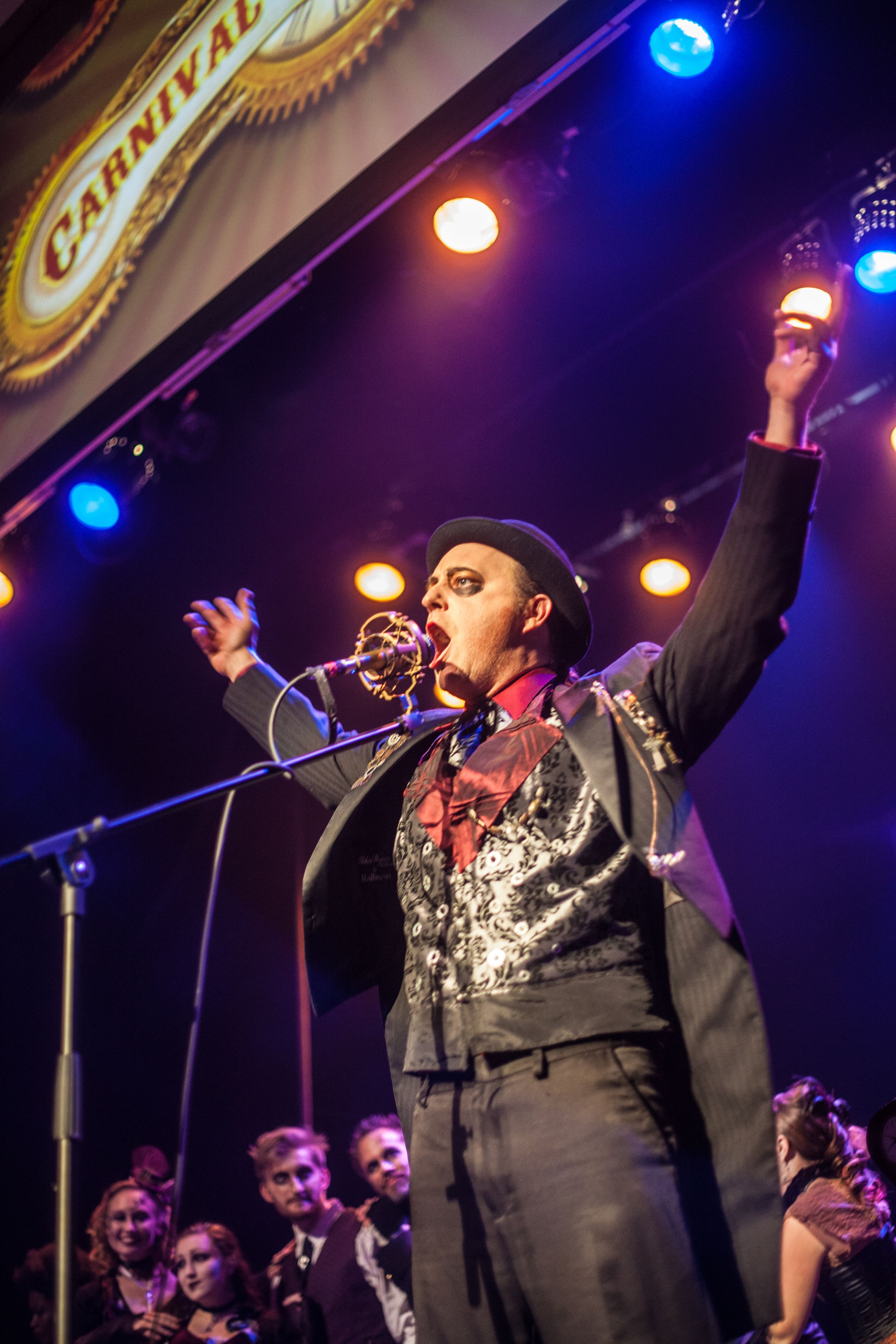Performer in black suit and hat singing on stage under colorful spotlights.