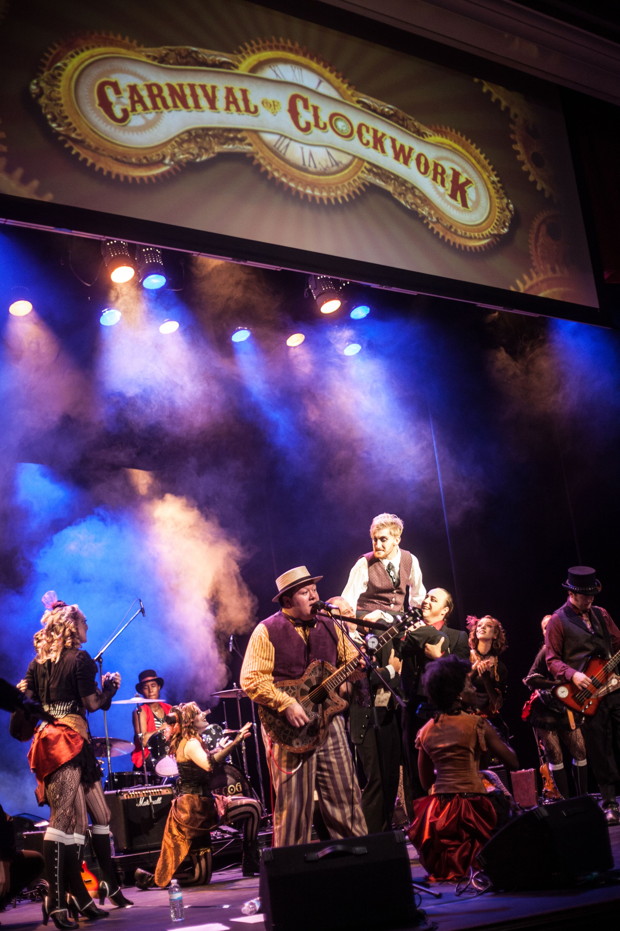 Band performs on stage under vintage 'Carnival of Clockwork' sign with dramatic blue and red theatrical lighting.