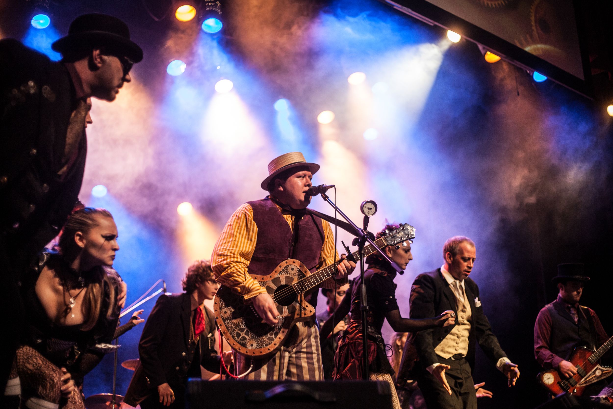 Steampunk-themed musical group performs on stage with vibrant blue lighting and fog effects.