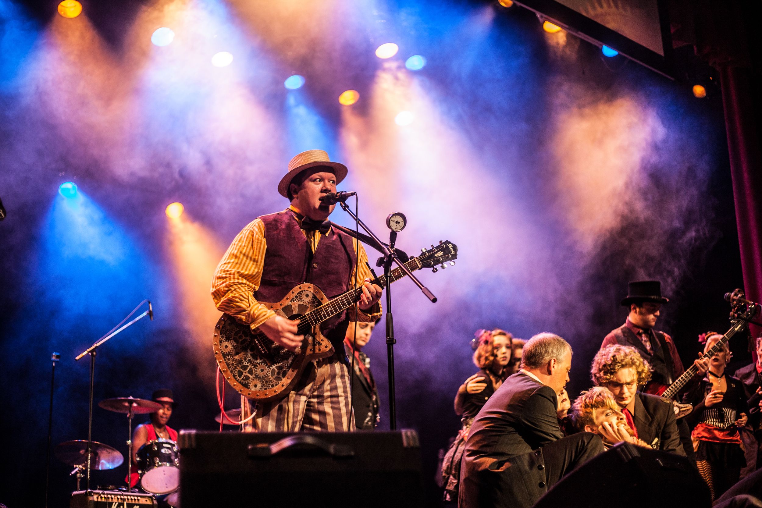 Musician in vintage attire plays guitar on stage with theatrical lighting and full band backdrop.