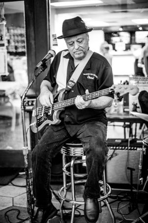 Bass player in black hat performs solo on stool in casual street setting.