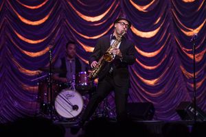 A saxophone player performs on stage with dramatic orange curtains and drum kit in the background.