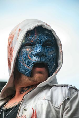 Person wearing a blue face mask and white hood in an artistic horror-style portrait.