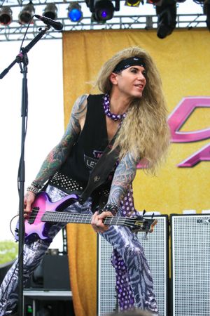 Glam rock bassist with long blonde hair performs on stage with pink guitar and elaborate costume.