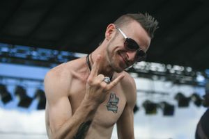 Shirtless performer gesturing on outdoor stage under bright daylight.