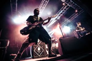 Energetic musician plays electric guitar on stage with dramatic backlighting and stage rigging visible.