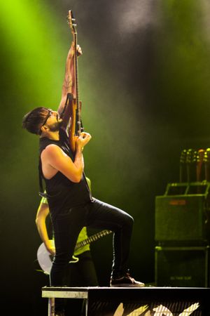 Musician performs energetically on stage with guitar raised upward against green lighting background.