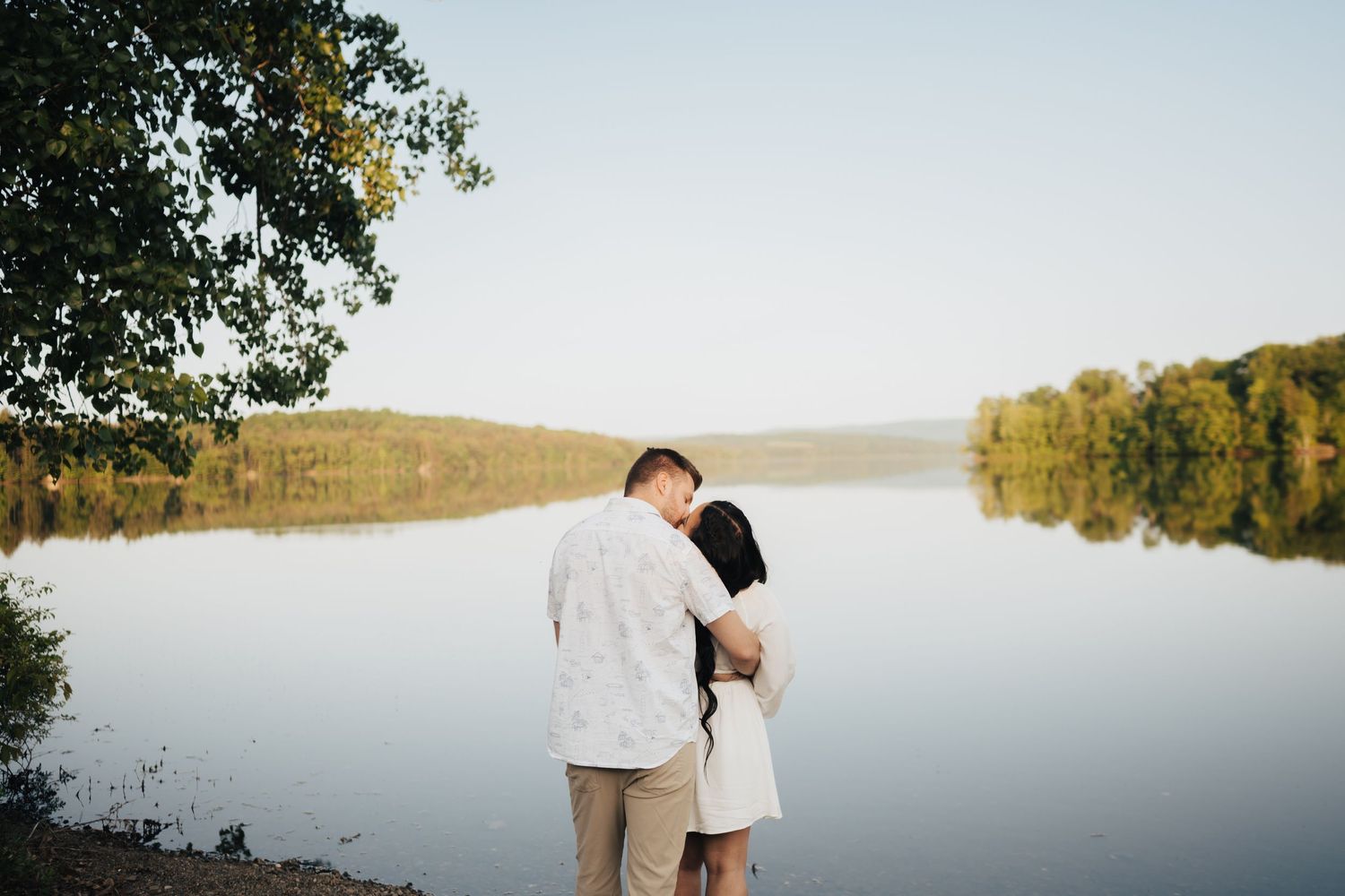 Romantic couple sharing a gentle kiss by a serene lake with lush green tree reflections.