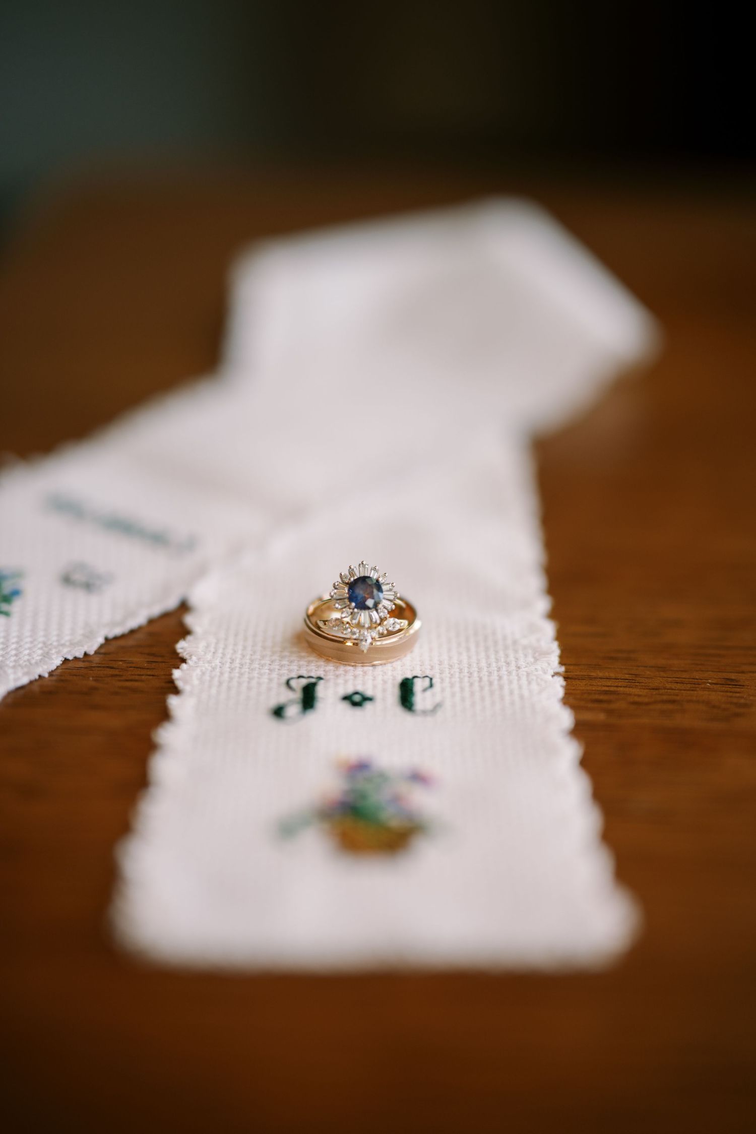 A sparkling engagement ring with a center stone rests on an embroidered white cloth with monogrammed initials.