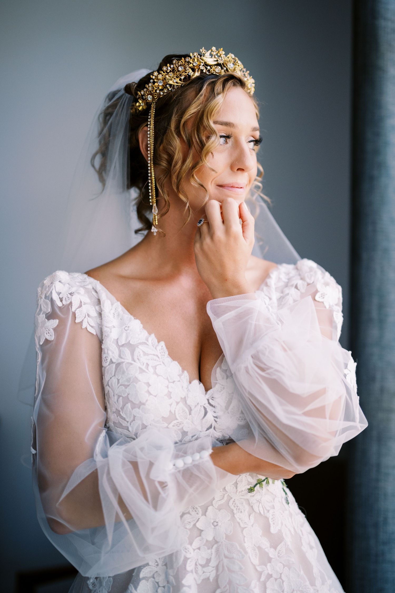 An elegant bride poses in a flowing white lace wedding gown with sheer sleeves and a gold ornate headpiece.