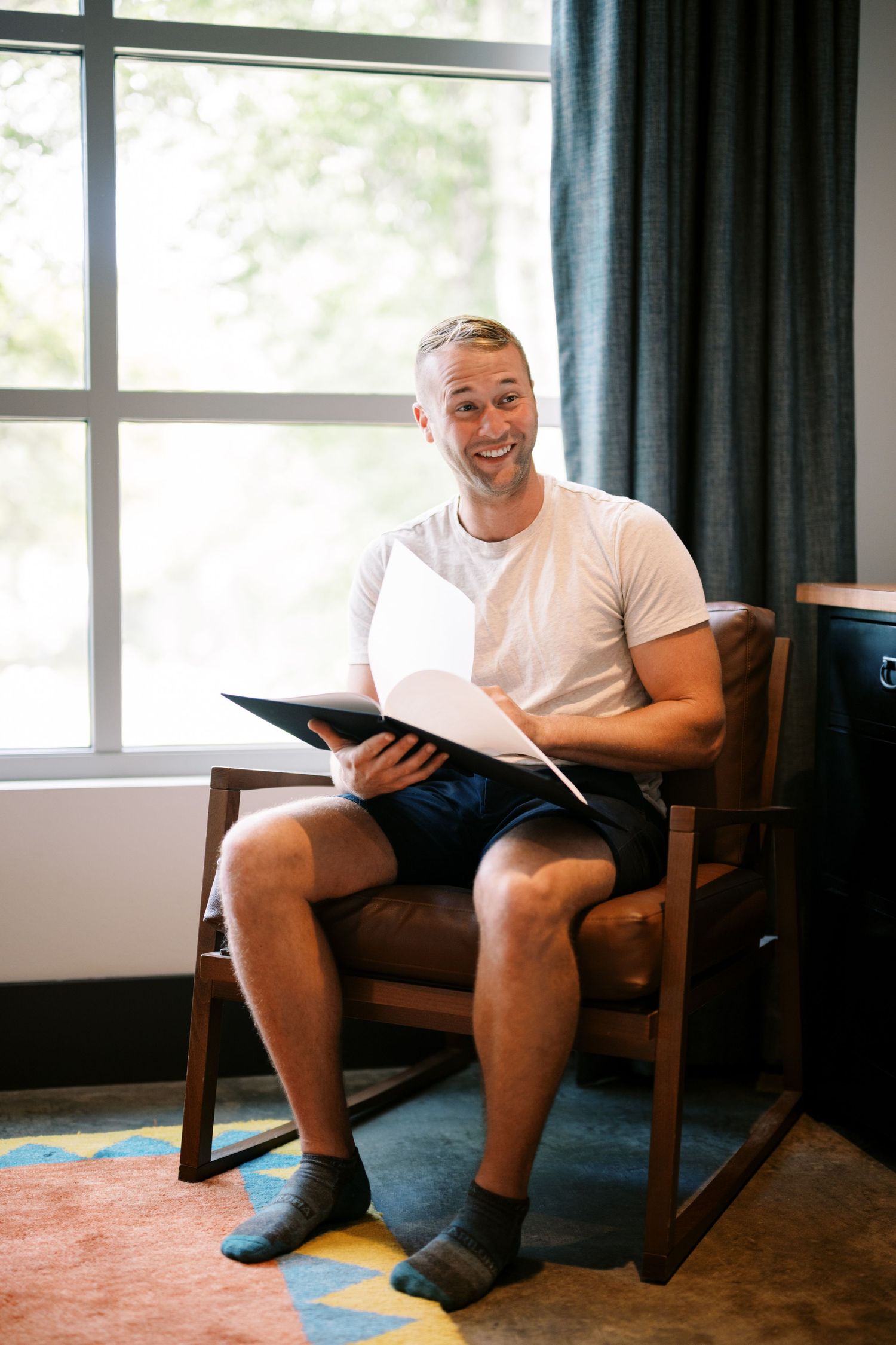 Person sitting in a wooden chair near a window reading documents and smiling in a cozy room setting.