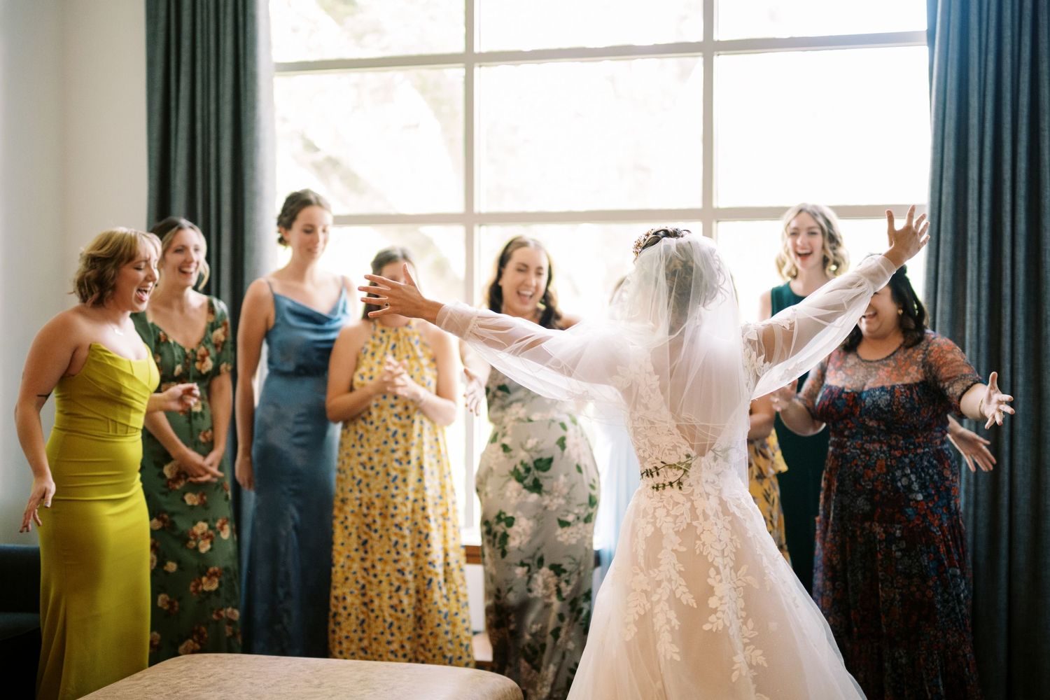 A bridal party helps adjust the bride's veil in front of a bright window while wearing colorful dresses.