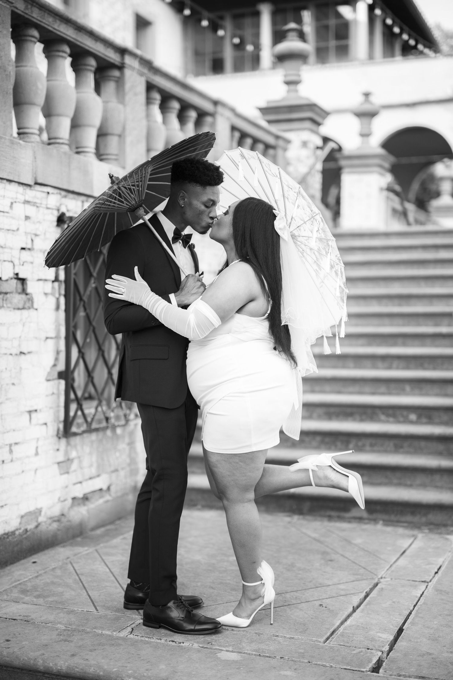 A romantic couple holding parasols embraces on brick steps of a grand building in black and white photography.