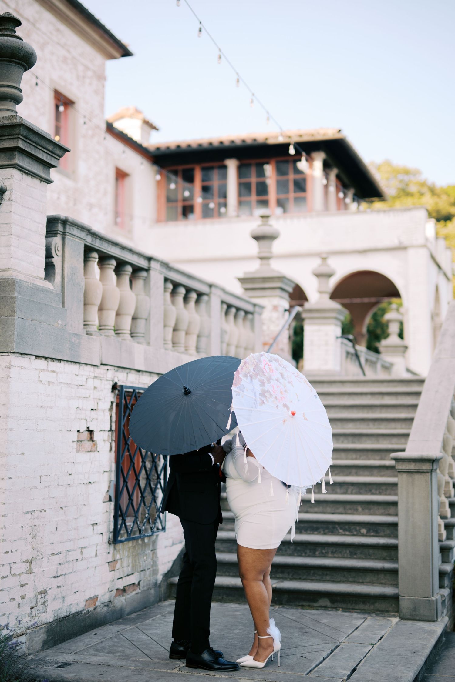 Two figures with contrasting black and white parasols share an intimate moment on historic stone steps.