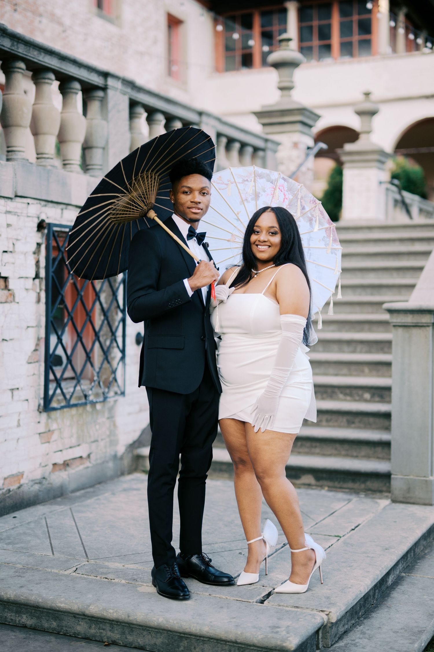 Couple poses elegantly on stone steps holding decorative parasols against classic architecture backdrop.