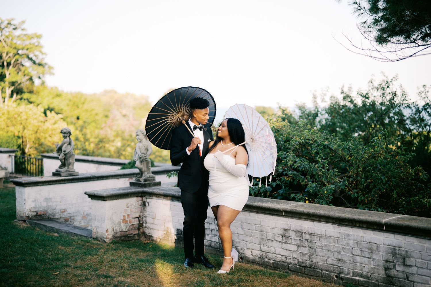 A couple in formal attire holding decorative umbrellas pose together in a garden setting during sunset.