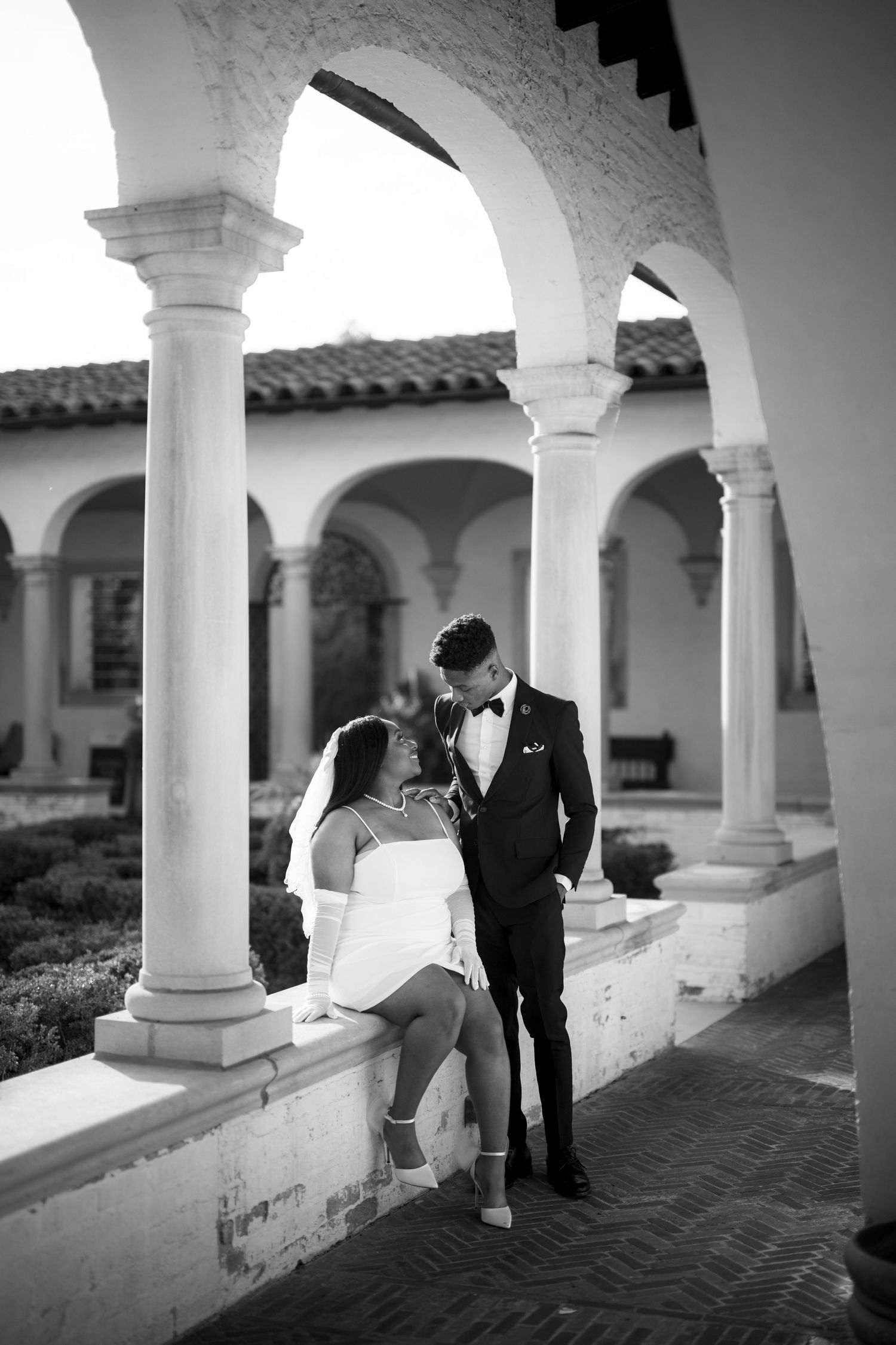 A romantic black and white photo of a couple in formal attire standing under Mediterranean-style arches and columns.