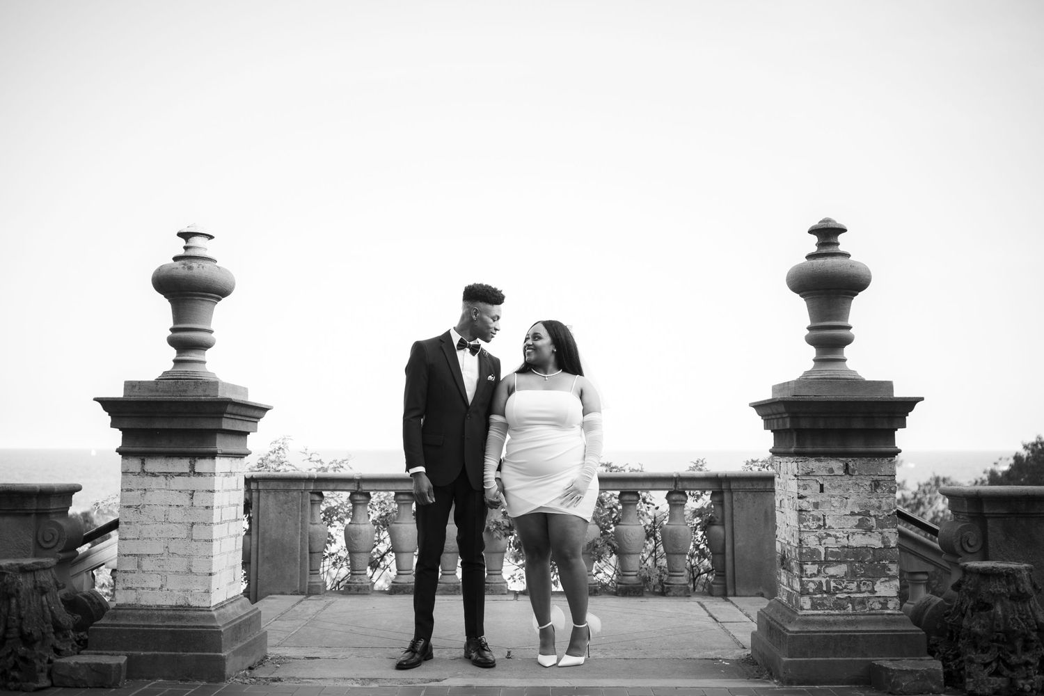 Black and white romantic photo of a couple standing between stone pillars on an outdoor terrace.