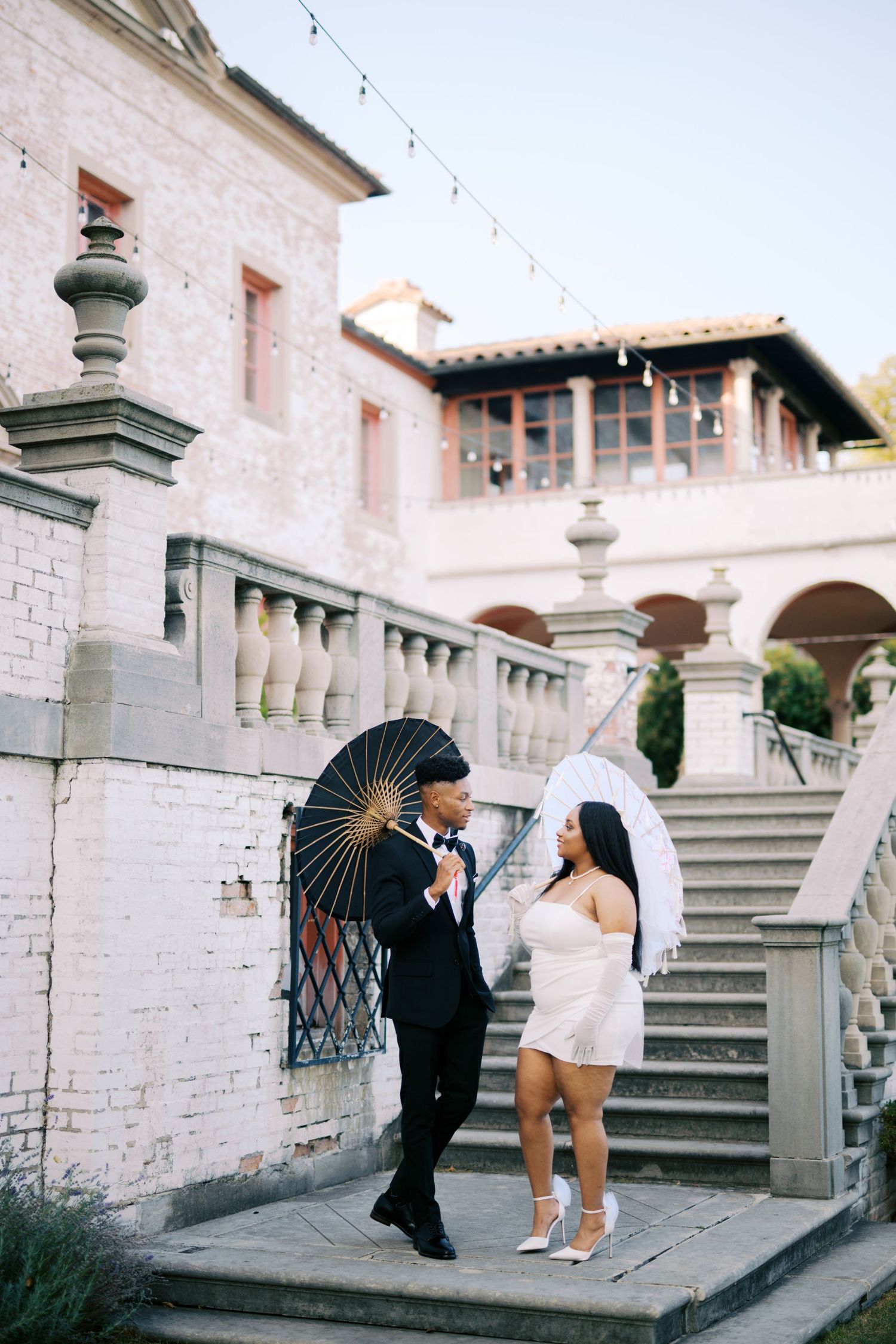 A couple with parasols stands on stone steps of a Mediterranean style villa during a romantic photoshoot.