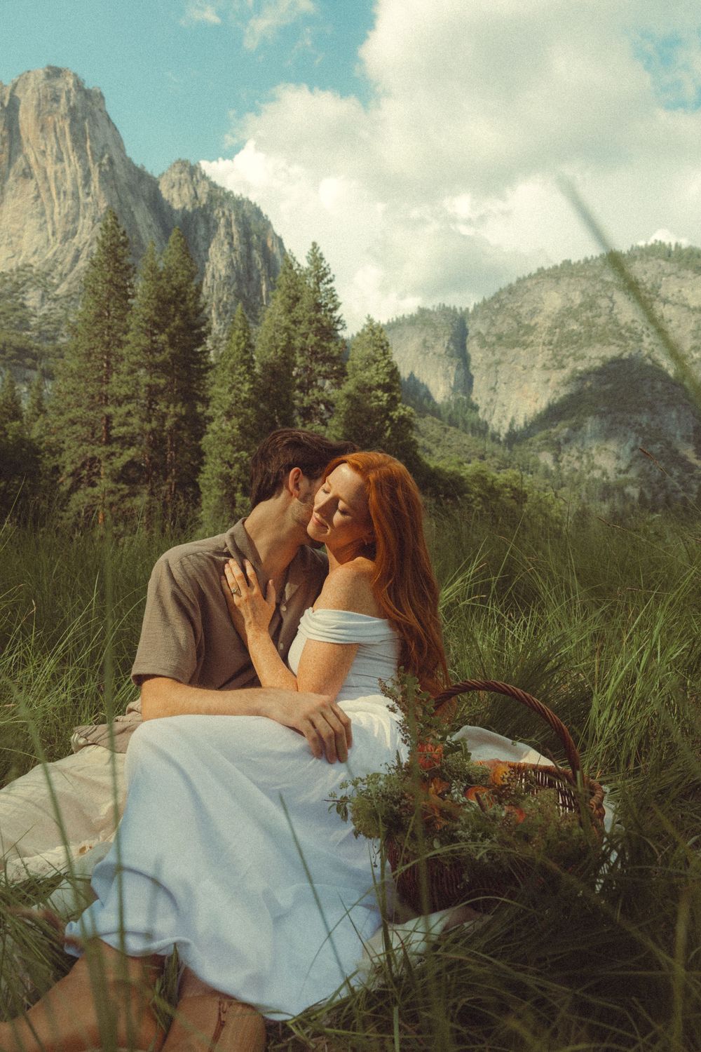 A romantic couple sits on a blanket in a meadow with Yosemite's majestic El Capitan mountain peaks in the background.