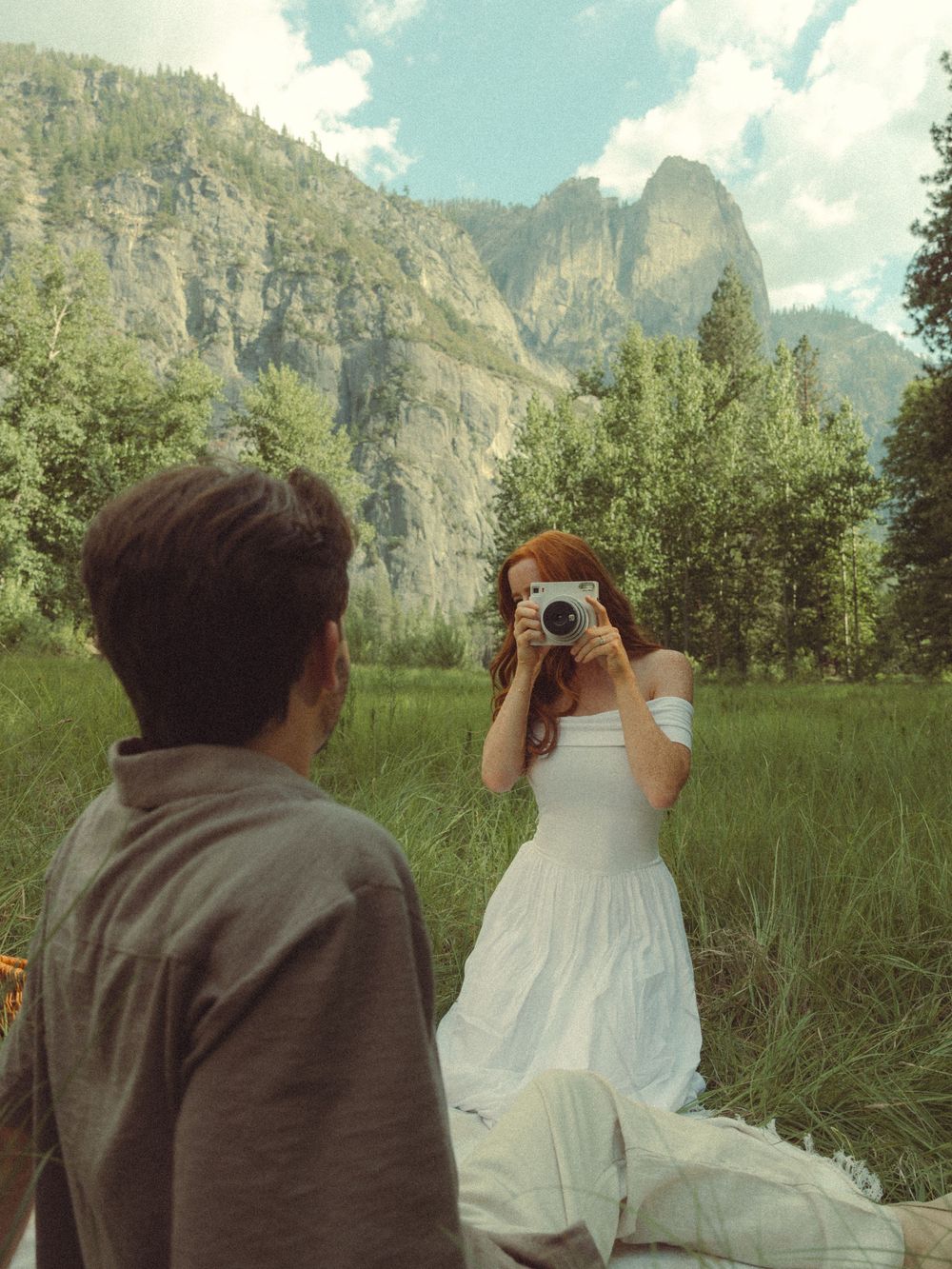 A photographer in a white dress captures photos with mountains and cliffs of Yosemite Valley in the background.