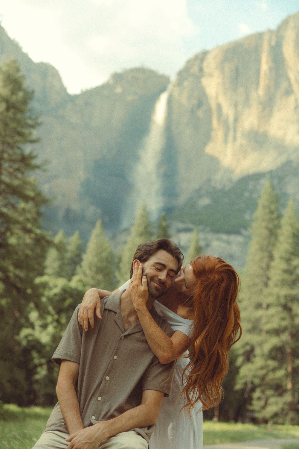 A romantic couple embraces in front of the majestic Yosemite Falls waterfall and granite cliffs on a sunny day.