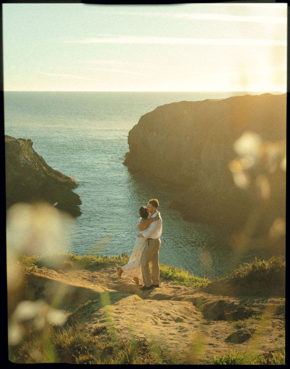 Couple embraces on coastal cliffs at sunset overlooking the ocean with golden light filtering through the scene.