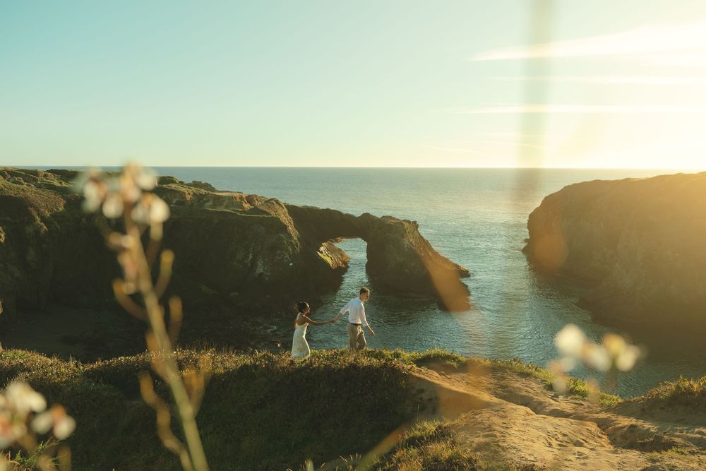 A silhouetted figure stands on coastal cliffs at sunset with ocean waves crashing against rocky arches in the background.