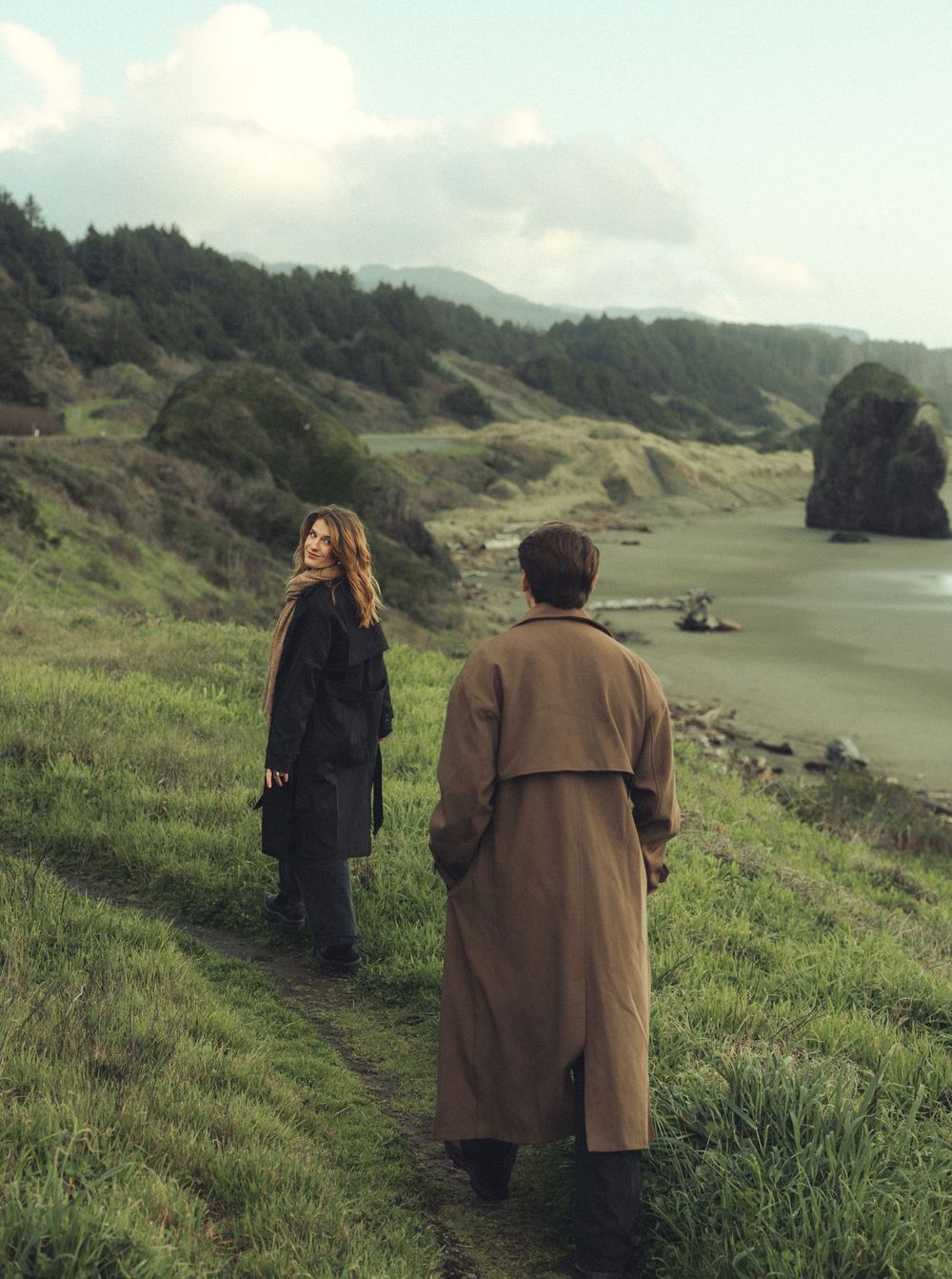 Two people in long coats walk along a scenic coastal path with green hills and dramatic rock formations by the water.