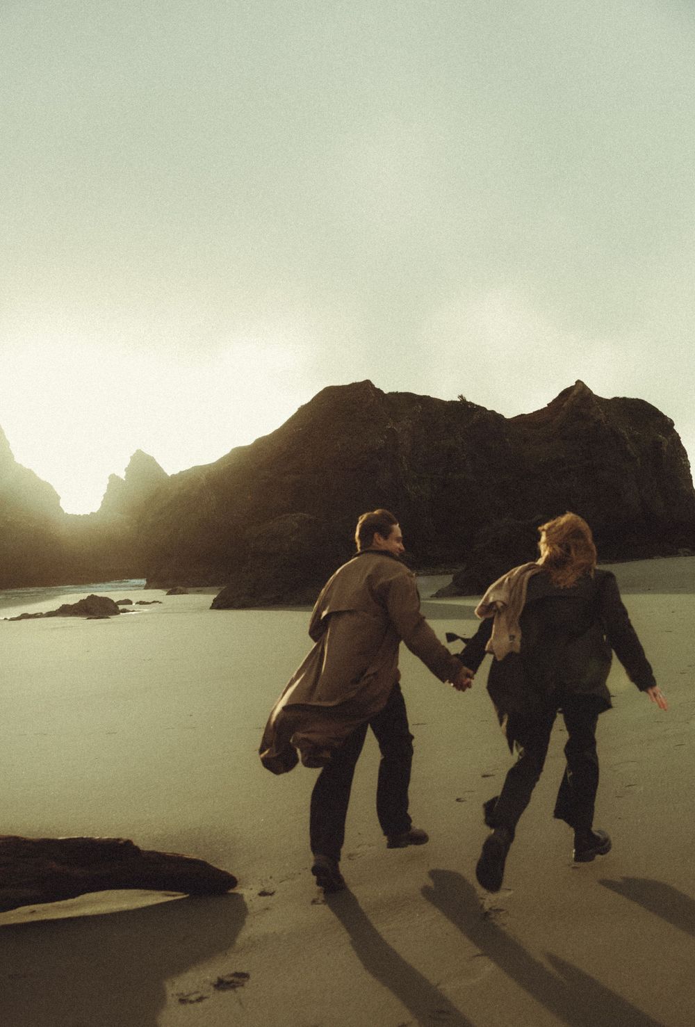 Two people hold hands and dance together on a misty beach at sunset with mountains silhouetted in the background.