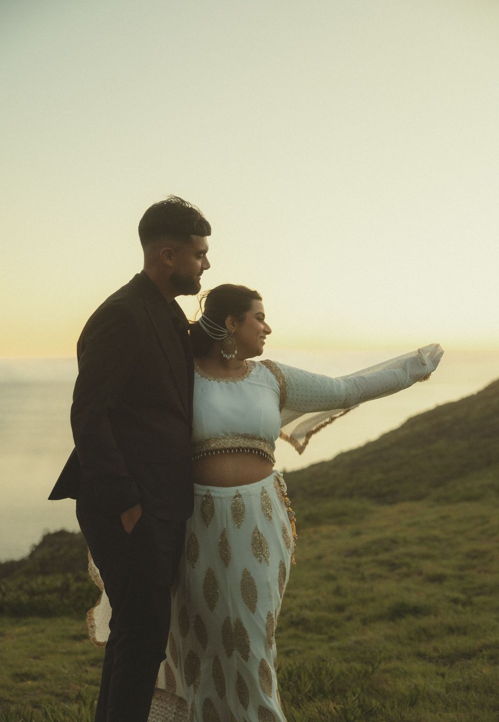 A couple stands on a grassy hillside at sunset, with one person pointing towards the horizon while the other embraces them.