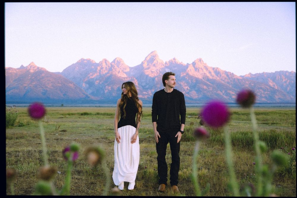 Couple standing in a mountain meadow with purple wildflowers, Grand Teton peaks glowing pink at sunset behind them.
