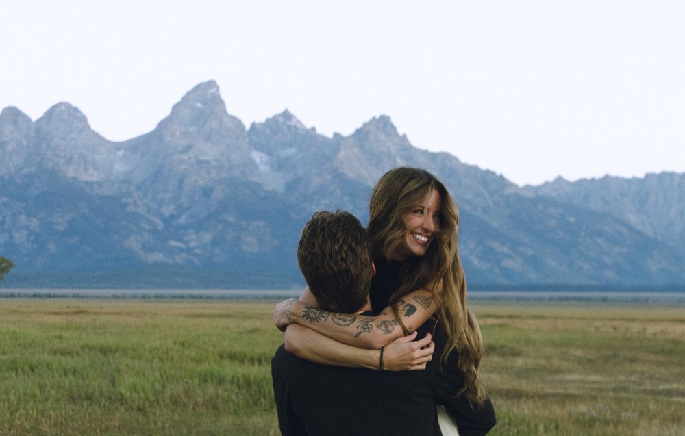 Couple embracing and laughing in a green meadow with the Grand Teton mountain range in the background.