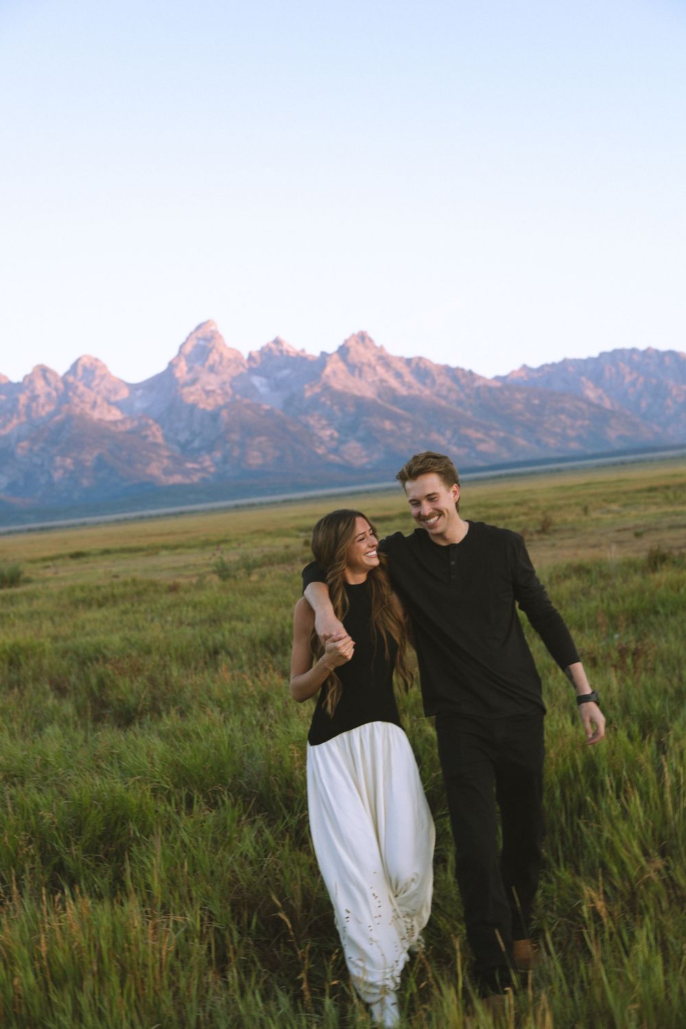 A smiling couple walks through a green meadow with the Grand Teton mountain range glowing pink at sunset behind them.