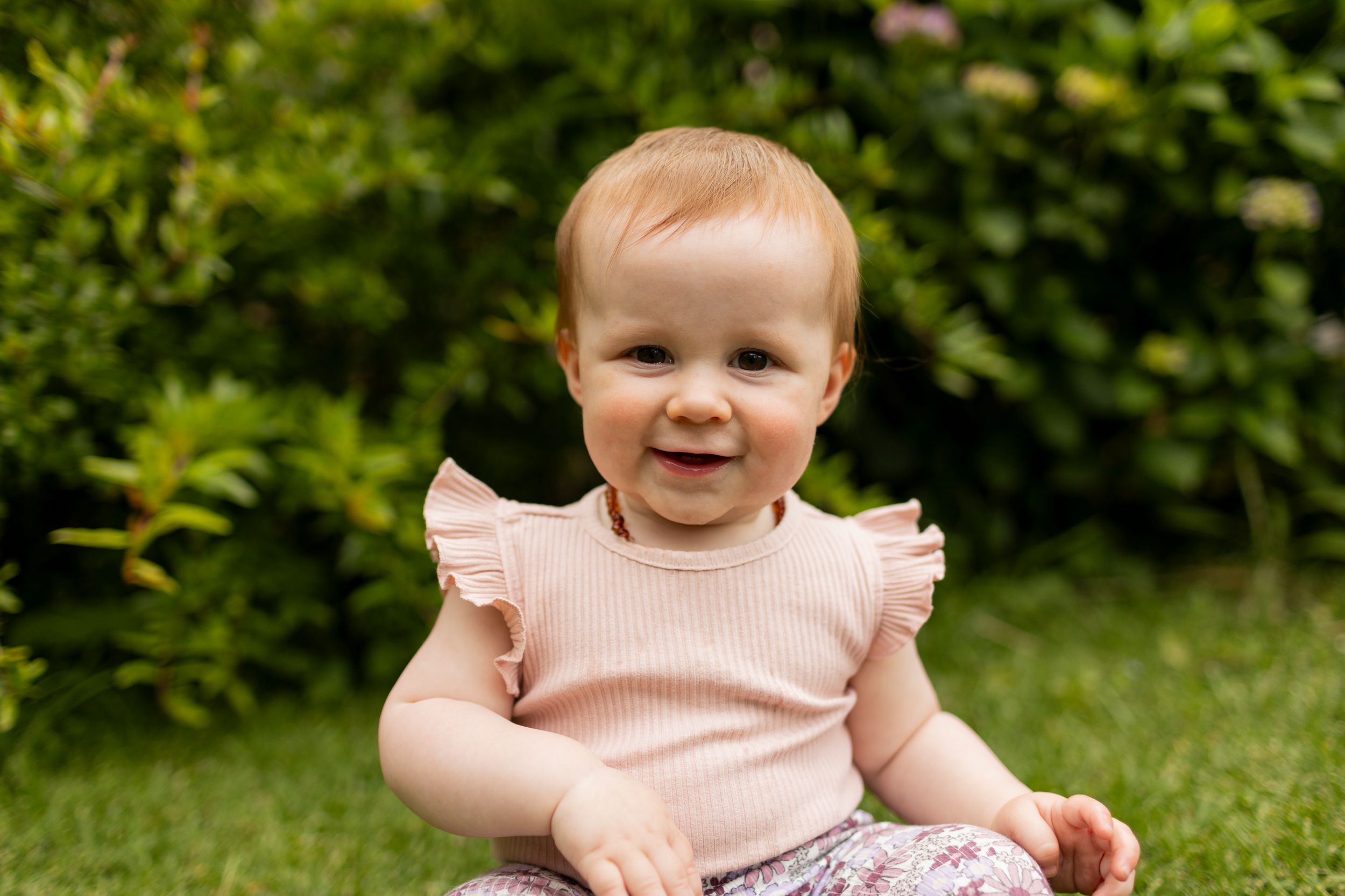 Small child wearing a pink ruffled top sits smiling in a green garden setting.