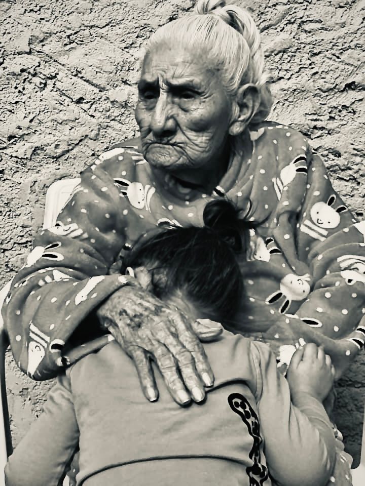 Elderly woman in patterned clothing embracing someone in black and white portrait.