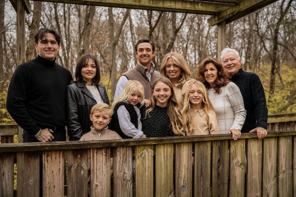 Multi-generational family wearing coordinated neutral colors poses together on a wooden deck in autumn woods.