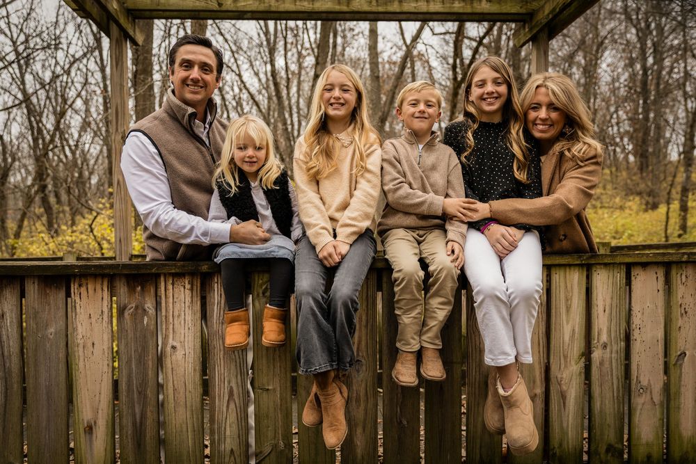 A family sits together on a rustic wooden deck railing during an autumn afternoon in casual coordinated outfits.