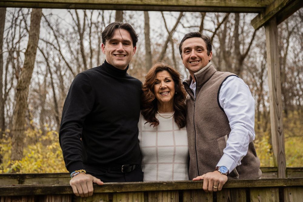 A group of people in winter clothing pose together on a rustic wooden bridge in a wooded area.