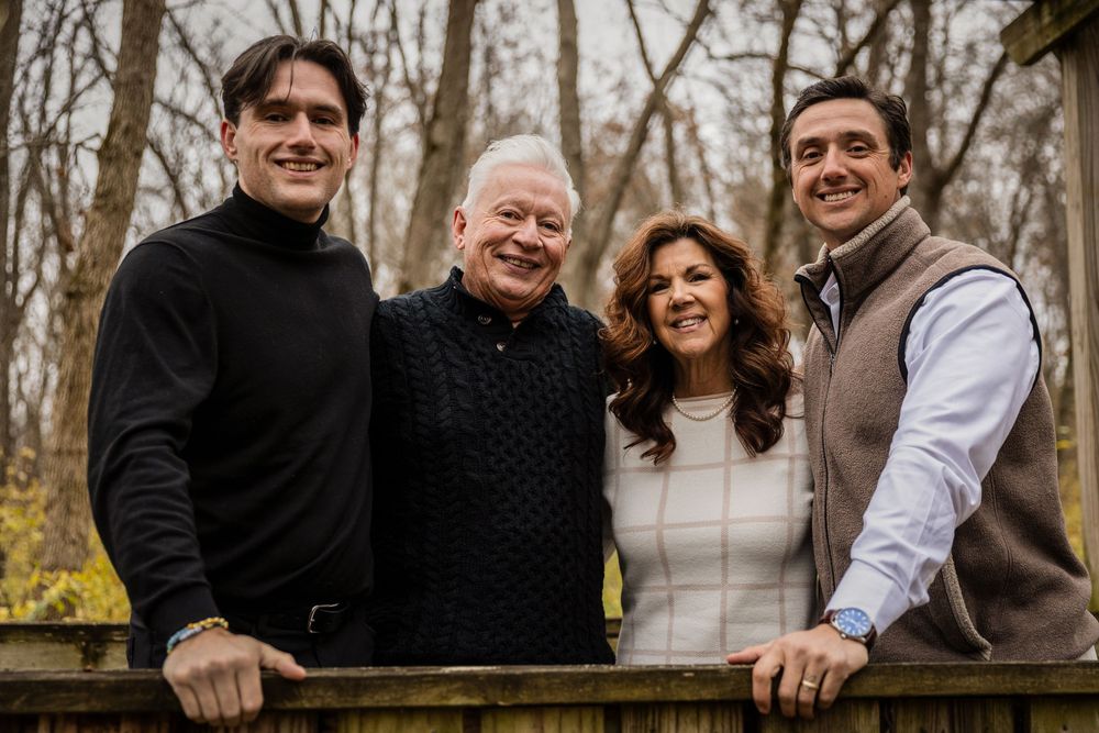 A group poses together outdoors in winter wearing dark coats and casual attire in a wooded setting.