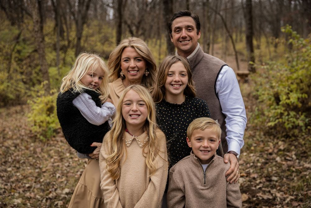A group of people posing together outdoors in a wooded area during autumn with fallen leaves on the ground.