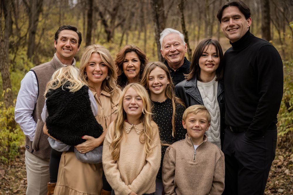 Family group wearing earth-toned sweaters and casual attire poses outdoors against fall foliage background.