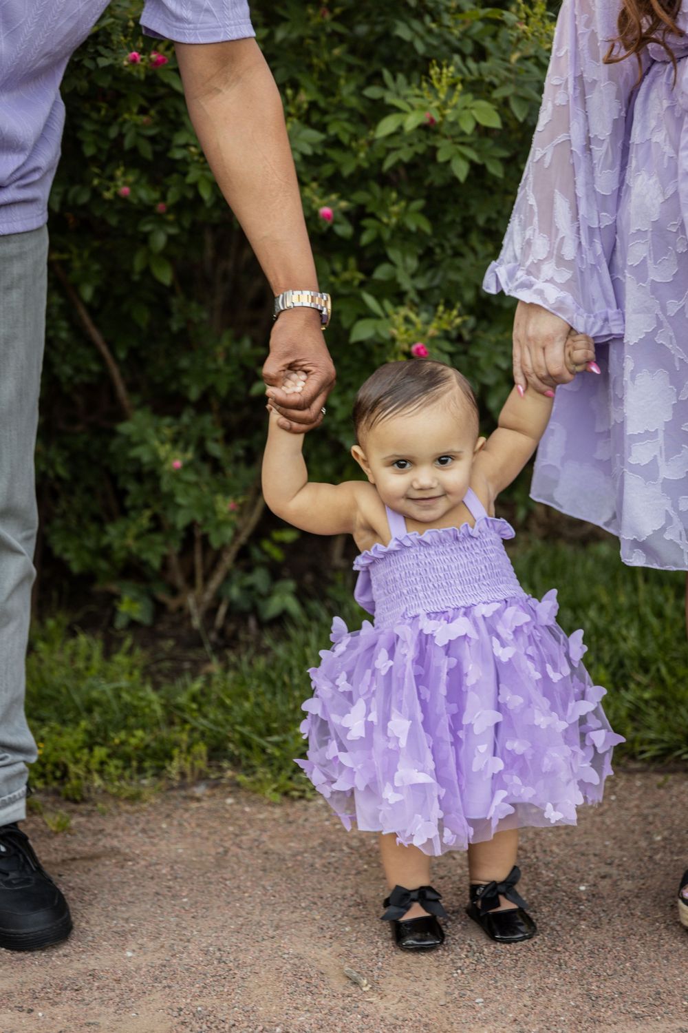 Toddler wearing a lavender tulle dress holds hands with adults in a garden setting.