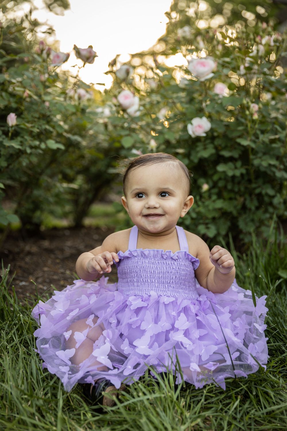 Infant wearing lavender tulle dress plays in grass near flowering bushes.