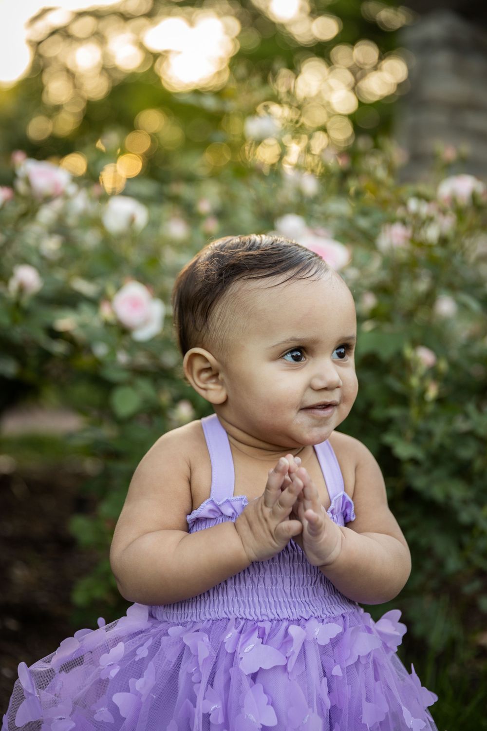 Baby in a ruffled purple dress smiling and clapping hands in a garden.