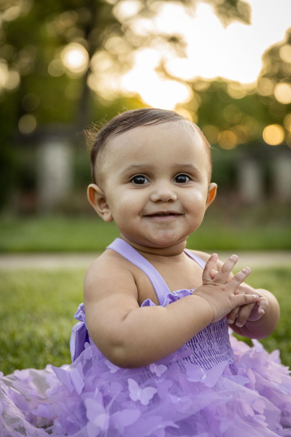 Smiling baby in purple dress poses against blurred garden background.