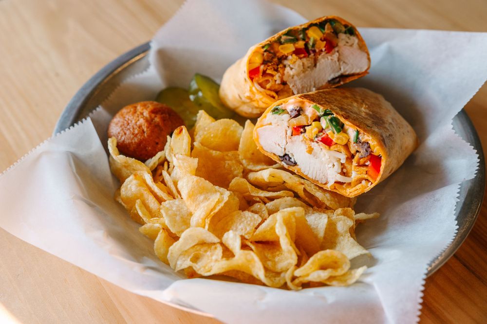 Wrapped sandwich filled with colorful ingredients served alongside crispy potato chips on white paper.